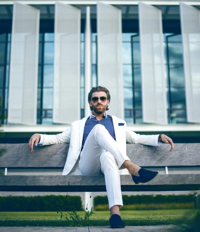 Hombre con aspecto exitoso, vestido con traje blanco y anteojos de sol, sentado frente a un edificio, representando ambición y éxito en ventas
